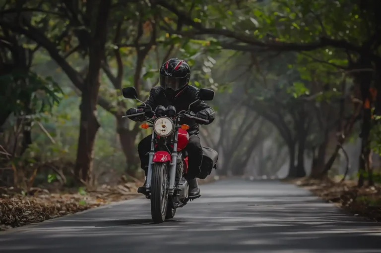 Um motociclista com sua moto de baixa cilindrada vermelha em uma rua com várias árvores no entorno
